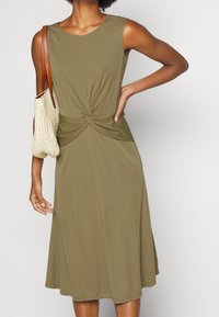 Oliwkowa, bezrękawnikowa sukienka z detalem w formie węzła w talii. Wykonana z miękkiego materiału, o luźnej sylwetce w kształcie litery A.
