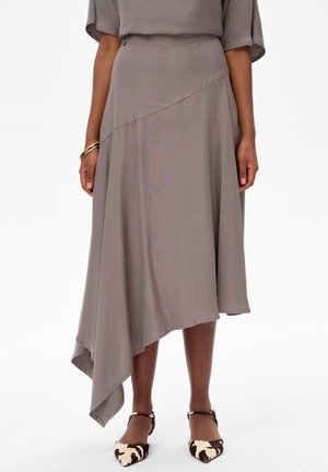 Kobieta w taupe asymetrycznej spódnicy midi z karbowanym dołem oraz w zwierzęcym wzorze na pasku dookoła kostki, nosząca szpiczaste baleriny.