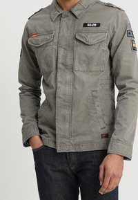 Grå militærjakke i stil med to brystlommer, knaplukning, skulderepauleter og patch-detaljer. Fremstillet af et letvægtigt, tekstureret stof.