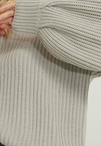Maglione grigio chiaro con texture a costine, caratterizzato da maniche ampie e arricciate e una vestibilità rilassata. Il materiale appare morbido e lavorato a maglia.