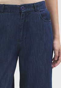 Pantaloni di jeans scuri con una texture liscia, dotati di un bottone viola, tasche anteriori e una vestibilità dritta.