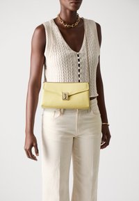 Borsa a tracolla gialla in pelle con una texture liscia, dotata di chiusura a patta e hardware metallico, abbinata a un top in maglia crema e pantaloni chiari.