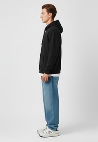 Felpa nera con tasca frontale, abbinata a jeans azzurri larghi e scarpe bianche con accenti neri. Design semplice, senza fantasie.