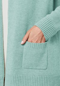 Hellblauer Cardigan aus weichem Strickmaterial, mit gerippten Bündchen und einer vorderen Tasche in klarer, rechteckiger Form.