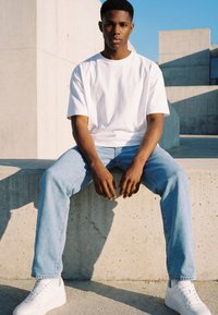 Jeune homme en t-shirt blanc, jean bleu clair et baskets blanches assis sur un rebord en béton devant un bâtiment moderne en béton.