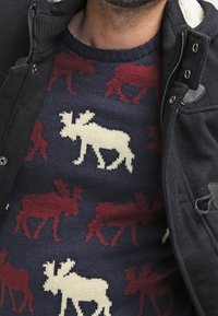 Maglione blu marino con motivi di alci color crema e bordeaux, tessuto a maglia texturizzato, collo rotondo, indossato sotto a una giacca nera con colletto in sherpa.