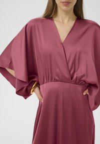 Rosa Satin-Kleid mit tiefem V-Ausschnitt, lockeren Fledermausärmeln und geraffter Taille. Mit einem goldenen Armreif am Handgelenk.