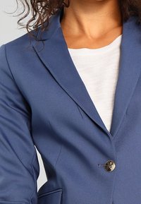 Námořnický modrý blazer s klopami, jedním knoflíkem a hladkým povrchem. Má dvě boční kapsy a přiléhavý střih.