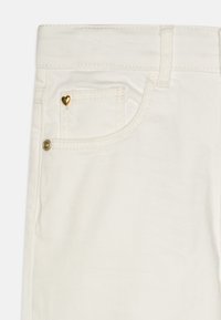 Valged denim püksid, mille esikoti peal on väike kuldne südame kaunistus, klassikalise viie tasku stiili ja vöödööbidega.