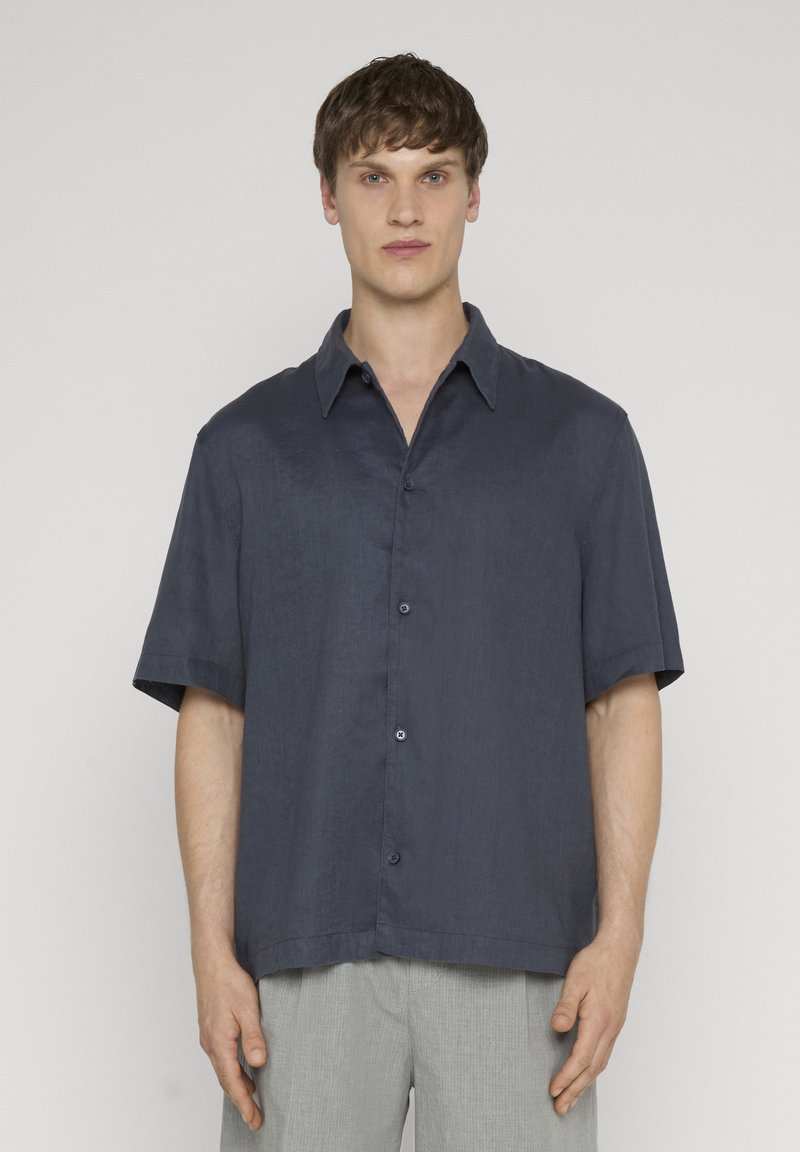 Homme portant une chemise bleu marine à manches courtes boutonnée et un pantalon gris clair, debout devant un fond uni gris clair.