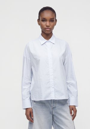 Femme à la peau foncée portant une chemise blanche à manches longues avec de fines rayures verticales et un jean bleu clair déchiré, debout dans une position neutre.