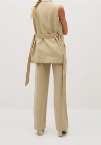 Beżowa kamizelka o dopasowanym kroju z wiązaniem w talii, posiada kieszenie boczne oraz prosty krój. Połączona z dopasowanymi spodniami o szerokich nogawkach i butami na słupku.