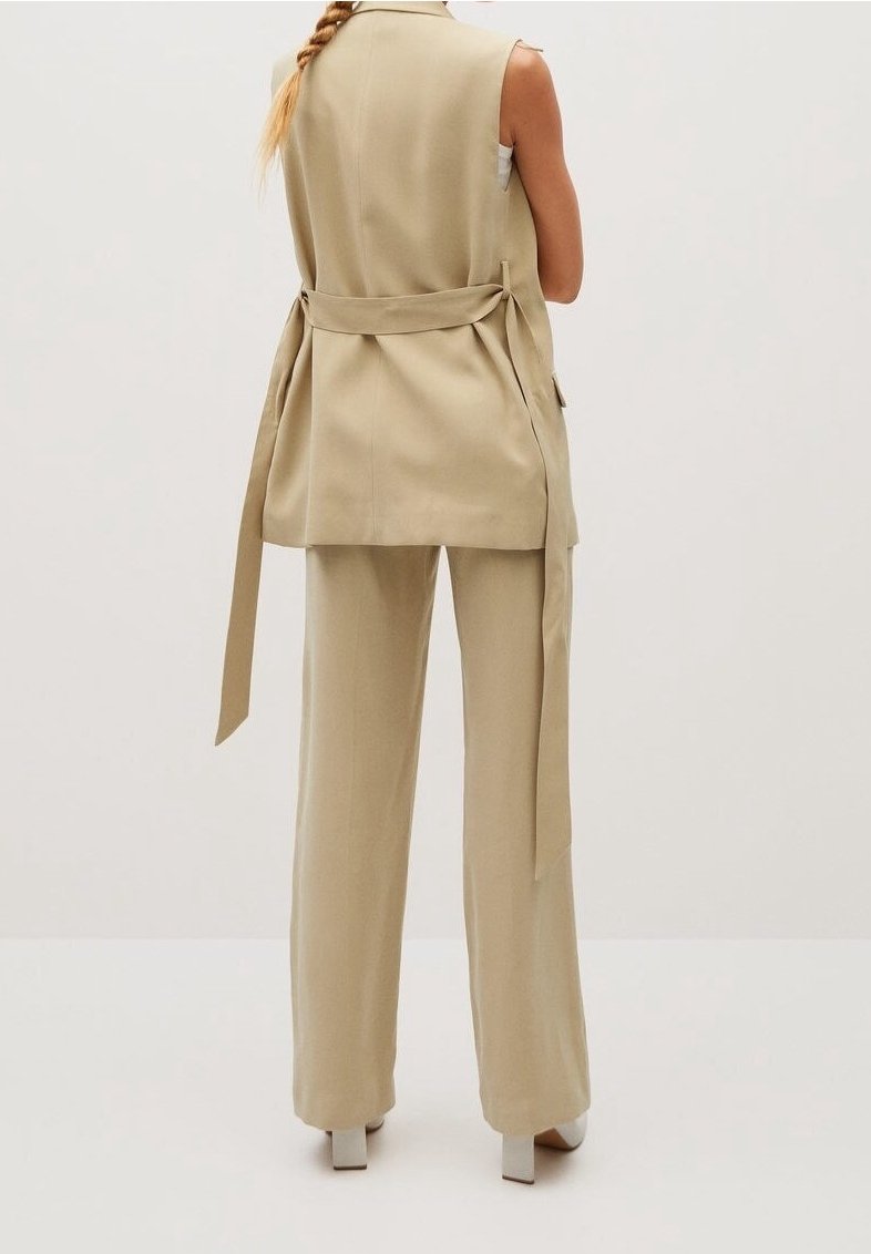 Beżowa kamizelka o dopasowanym kroju z wiązaniem w talii, posiada kieszenie boczne oraz prosty krój. Połączona z dopasowanymi spodniami o szerokich nogawkach i butami na słupku.