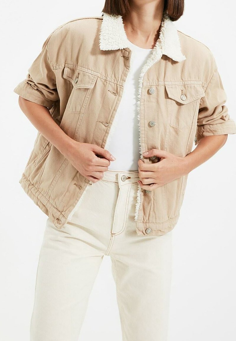 Béžová denim bunda s huňatým bílým límcem, předními kapsami a stříbrnými knoflíky. Nosená přes bílou blůzu a světlé džíny.