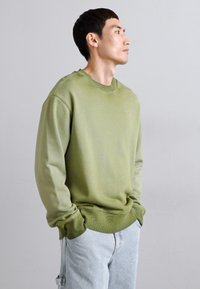 Sweatshirt girocollo verde oliva realizzato in tessuto morbido, con polsini e fondo a costine, indossato con jeans azzurri chiari. Taglio classico e design minimal.