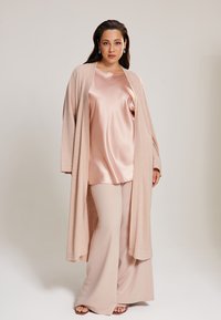 Langer, offener Cardigan in Hellbeige, getragen über einem seidigen, rosafarbenen Top und passenden, weit geschnittenen Hosen, die glatte Texturen und sanfte Farben präsentieren.