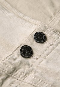 Beige stof met een ribbeltextuur heeft twee zwarte metalen knopen met een gegraveerd logo, zichtbare stikdetails en een subtiele glans.