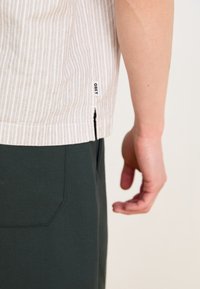Chemise à manches courtes rayée de couleur claire avec une étiquette, associée à un pantalon vert foncé. Présente des fentes latérales pour faciliter le mouvement.