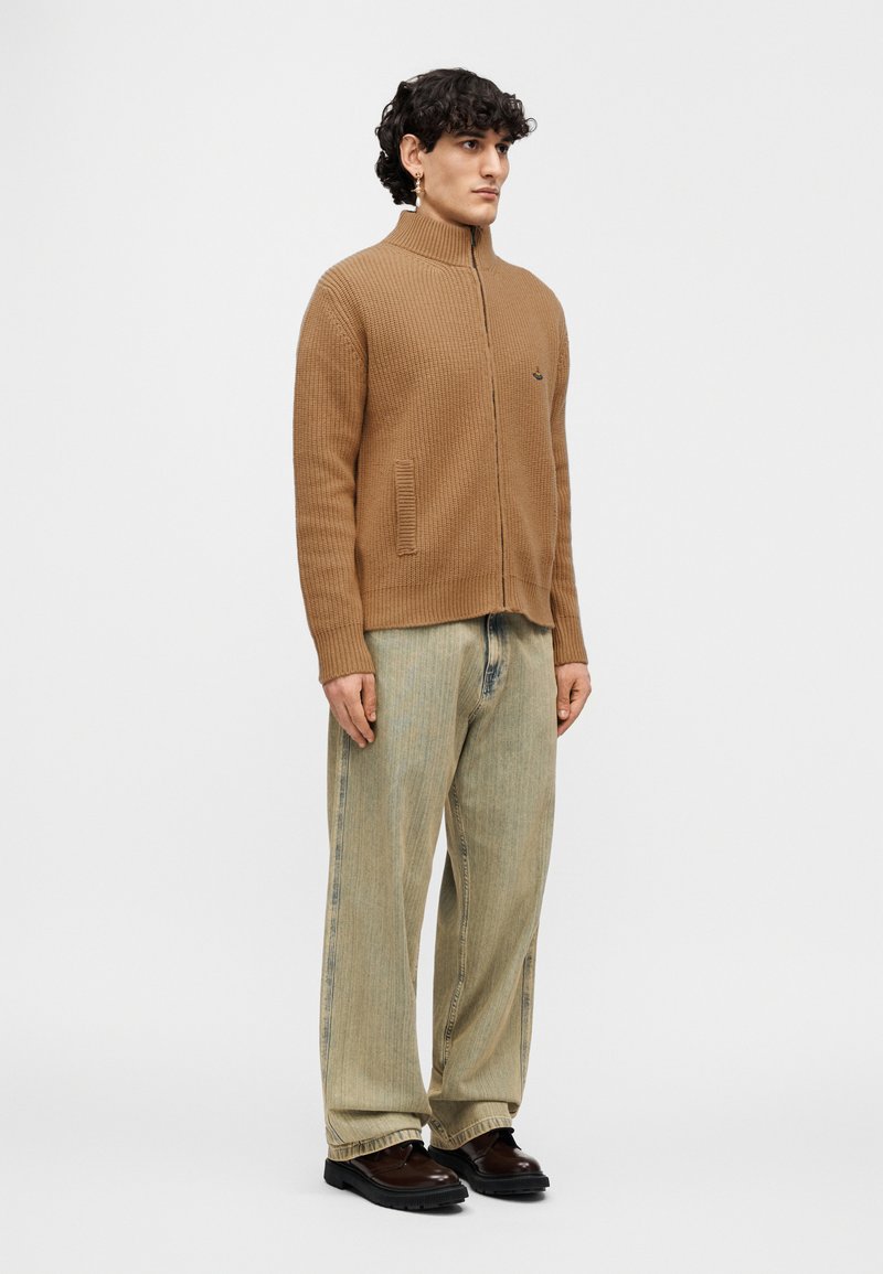 Brown ribbed cardigan with high collar, two front pockets, and a relaxed fit, paired with light blue wide-legged jeans and black shoes.