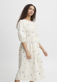 Robe en coton blanc avec broderies florales en rose, violet et vert. Corsage smocké, manches bouffantes et silhouette mi-longue en A.