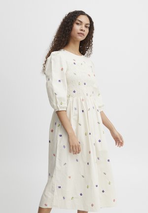 Robe en coton blanc avec broderies florales en rose, violet et vert. Corsage smocké, manches bouffantes et silhouette mi-longue en A.