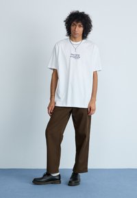 Vit oversized t-shirt med texten "the new originals jazz club" på bröstet, tillsammans med bruna avslappnade byxor och svarta skor.