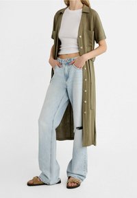Abito camicia leggero verde oliva con bottoni, abbinato a jeans a gamba larga azzurri con un ginocchio strappato. Sandali marroni completano il look.