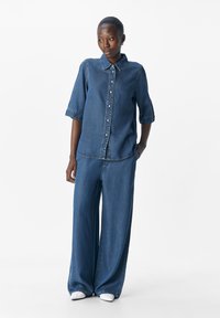 Mujer de pie vestida con una camisa de mezclilla azul de botones y mangas hasta los codos, combinada con unos vaqueros holgados a juego y zapatos blancos.