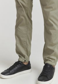 Khaki-väriset rento-funktiot pantaloonit, joissa on kapenevat lahkeet ja napin yksityiskohta, yhdistettynä mustiin nahkakenkiin, joissa on valkoinen kumipohja.