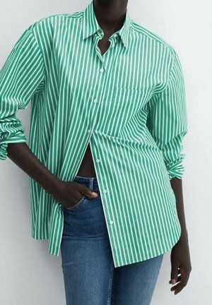 Personne portant une chemise à rayures verticales vertes et blanches avec boutons, manches retroussées et jean bleu, main dans la poche, sur fond uni.