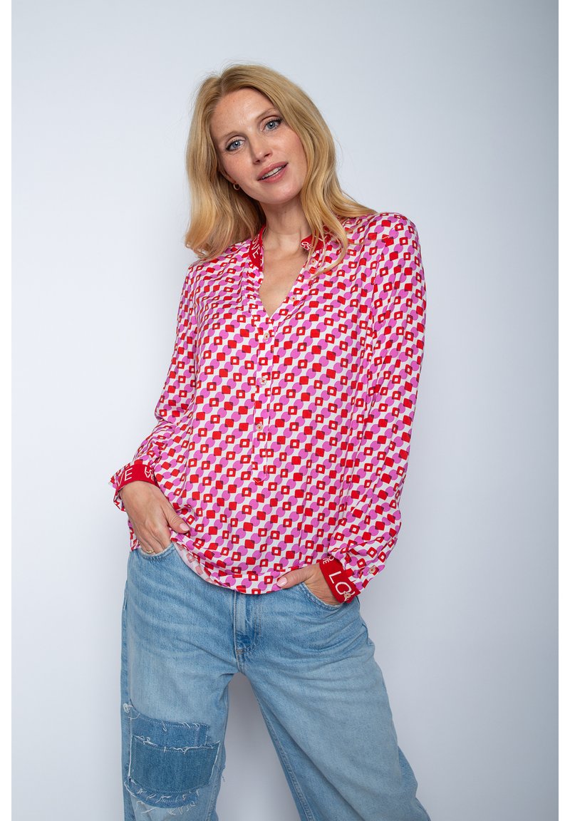 Rote und rosa gemusterte Bluse mit V-Ausschnitt, langen Ärmeln und "LOVE" Akzenten an den Manschetten. Getragen mit hellblauen, distressed Jeans.