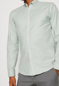 Jasnozielona koszula na guziki z długimi rękawami, wykonana z miękkiego materiału, z ostro zakończonym kołnierzykiem i subtelną fakturą. Zapięcie na guziki z przodu.