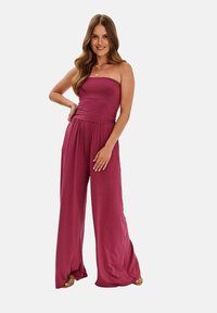 Burgundská bezramenná kombinéza z hladké tkaniny s volným střihem, širokými nohavicemi a nařaseným pasem. Obsahuje jemné drapování.