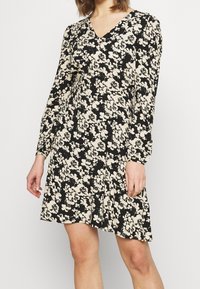 Kvinna i en svart långärmad V-ringad klänning med krämfärgad blommönster, stående med avslappnade armar vid sidorna mot en enkel bakgrund.