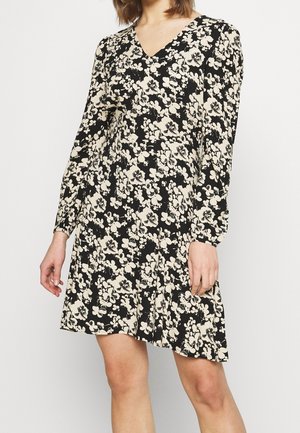 Vrouw in een zwart langetermijn V-halsjurk met crèmekleurige bloemenprint, staand met ontspannen armen langs de zijkanten tegen een effen achtergrond.
