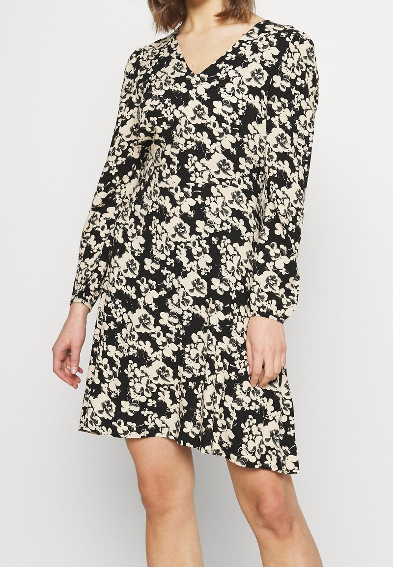 Kvinna i en svart långärmad V-ringad klänning med krämfärgad blommönster, stående med avslappnade armar vid sidorna mot en enkel bakgrund.