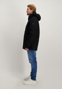 Zwarte parka met een capuchon, ritssluiting aan de voorkant en zijzakken, gecombineerd met blauwe jeans en witte sneakers, gepresenteerd vanuit een zijaanzicht.