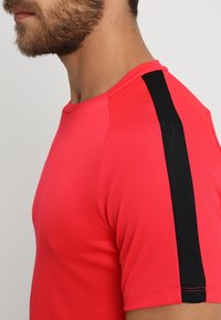 Punainen urheilupaita, jossa on musta pystysuora raita olkapäällä, valmistettu sileästä ja venyvästä kankaasta. Lähikuva sivuprofiilista.