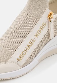 Sneaker slip-on beige lavorato a maglia con superiore texturizzato e inserzioni in pelle liscia, caratterizzato da un logo dorato e una suola in gomma bianca.
