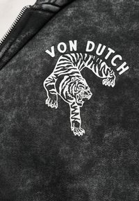 Σκούρο γκρι ύφασμα με λευκό γραφικό που απεικονίζει έναν τίγρη που ουρλιάζει και το κείμενο "VON DUTCH" από πάνω, ορατό φερμουάρ στη αριστερή πλευρά.