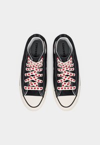 Ζευγάρι μαύρα αθλητικά παπούτσια Converse με λευκά καπάκια στα δάχτυλα και λευκά κορδόνια διακοσμημένα με  κόκκινες καρδιές, απεικονιζόμενα από πάνω σε λευκό φόντο.