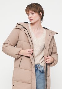 Joven vestido con una chaqueta de plumas beige sobre un suéter de punto crema y jeans azules, ajustando la cremallera de la chaqueta, mirando hacia un lado.