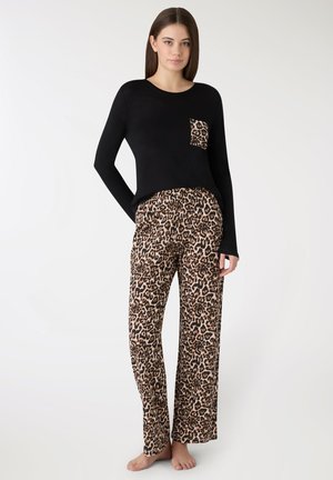 Donna in piedi a piedi nudi che indossa una maglia nera a maniche lunghe con tasca a stampa leopardata e pantaloni a gamba larga coordinati con stampa leopardata, su sfondo bianco.