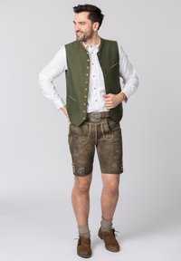 Gilet matelassé vert avec des accents dorés, chemise blanche, shorts en cuir marron avec un embossage floral, chaussettes grises et chaussures marron.
