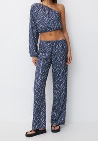 Modrý květinový dvoudílný outfit s jedním ramínkem, zahrnující krátký top s dlouhým rukávem a volné kalhoty. Materiál má jemnou strukturu.