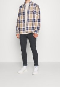 Camisa de cuadros en beige, azul marino y gris; camiseta gris debajo; jeans oscuros; zapatillas blancas con acentos en azul claro.