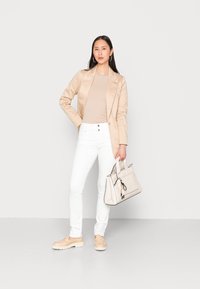 Blazer de satén beige sobre una blusa color tostado, jeans blancos ajustados y mocasines de un beige claro; sostiene un bolso estructurado color crema con acentos.