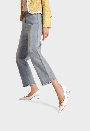 Tacchi a punta bianchi con un accento dorato, abbinati a jeans blu chiaro cropped con orli grezzi, indossati da un modello con una giacca gialla testurizzata.