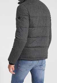 Chaqueta acolchada gris con cuello alto, diseño acolchado y un parche de logo negro en el hombro. Combinada con jeans azules.