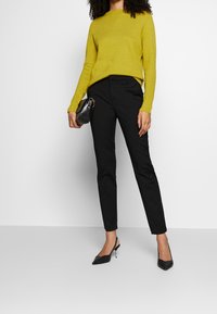 Femme portant un pull jaune à manches longues, un pantalon noir sur mesure, des escarpins noirs slingback, tenant un petit sac à main noir sur fond blanc.